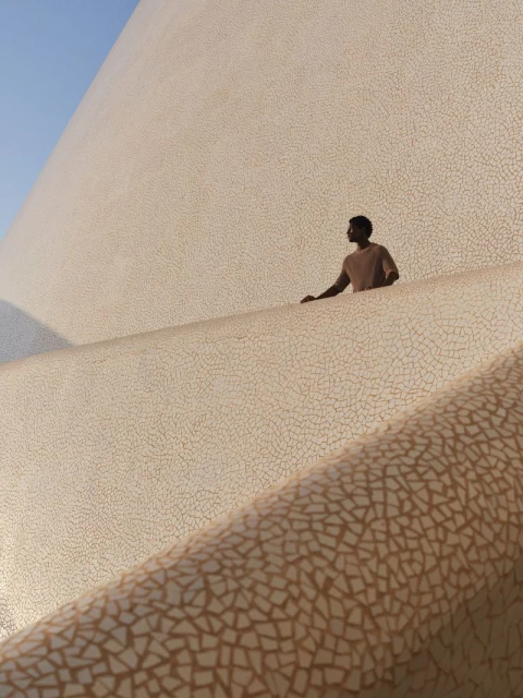 An African American man stands on the ridge of a large architectural structure with geometric, mosaic-like surface under a clear blue sky.