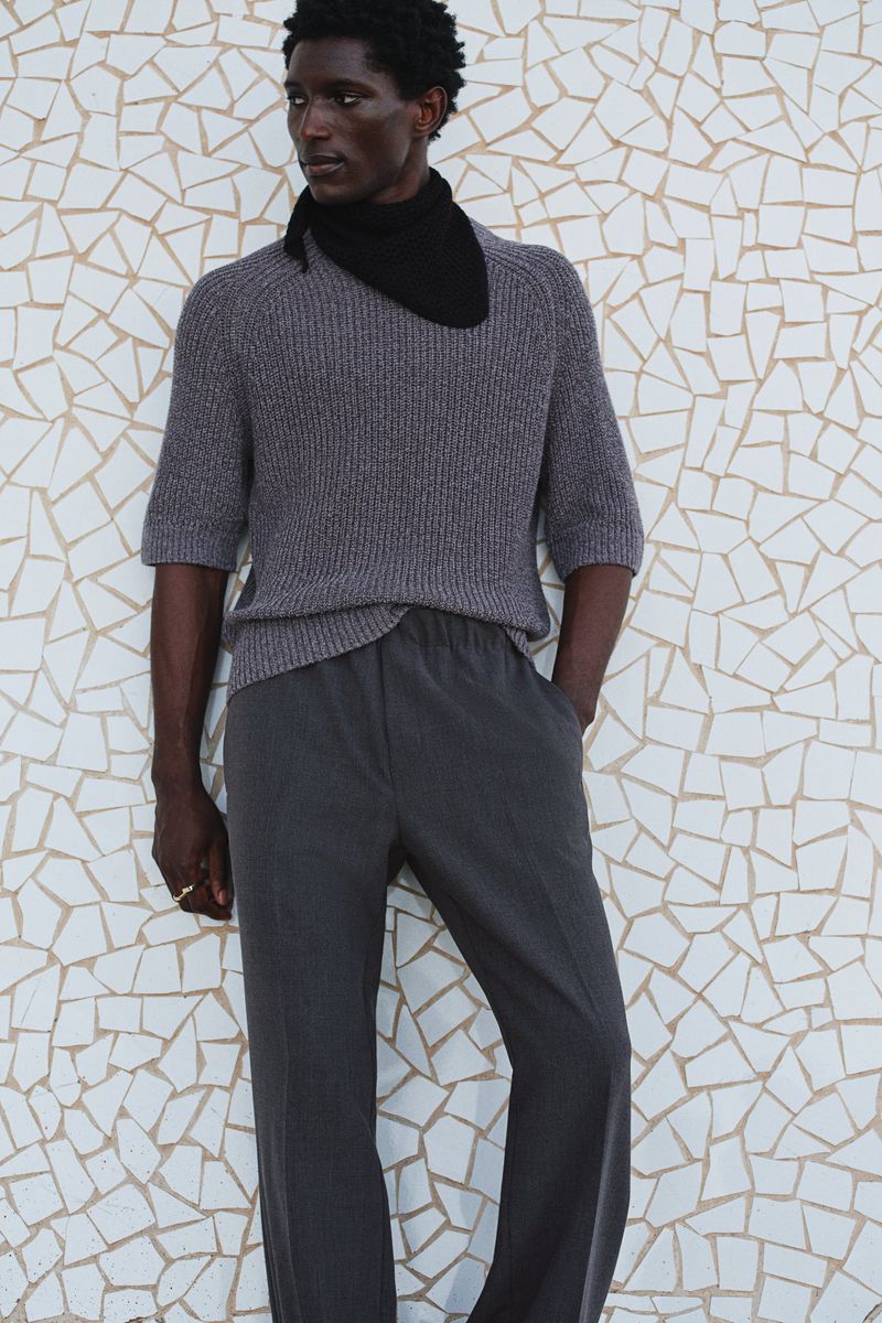Studio shot of a Black male model in casual gray knitwear, complemented by a black scarf, posing against a geometric tile wall.