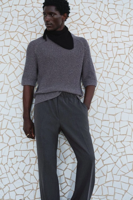 Studio shot of a Black male model in casual gray knitwear, complemented by a black scarf, posing against a geometric tile wall.