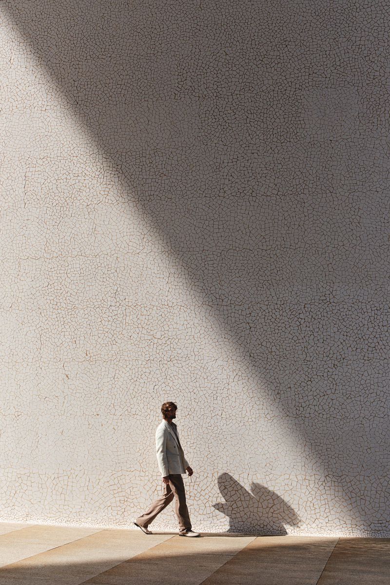 Full-length shot of a man walking in a suit alongside a sunlit wall with a cast shadow, creating a simple and elegant composition.