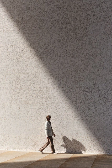Full-length shot of a man walking in a suit alongside a sunlit wall with a cast shadow, creating a simple and elegant composition.