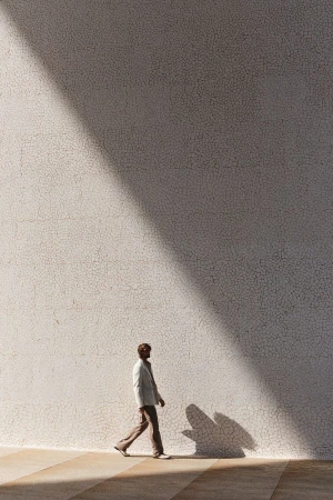 Full-length shot of a man walking in a suit alongside a sunlit wall with a cast shadow, creating a simple and elegant composition.