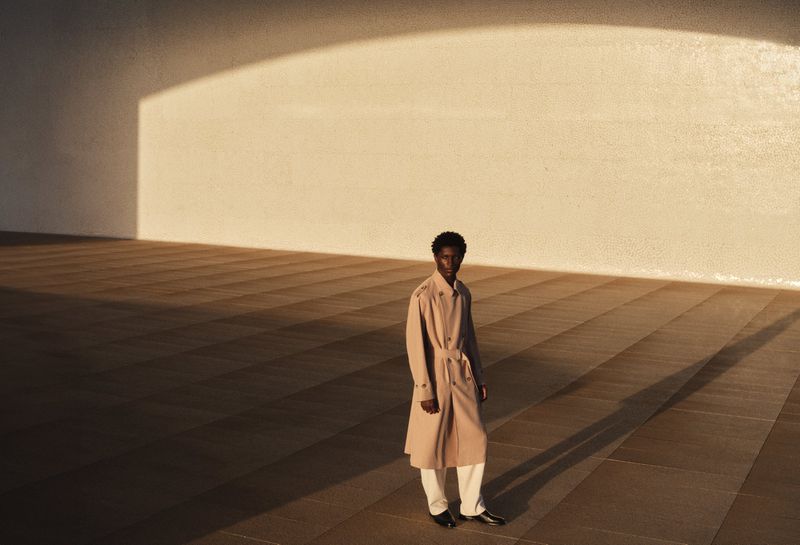 Model standing on a tiled surface wearing a beige trench coat under dramatic lighting for H&M