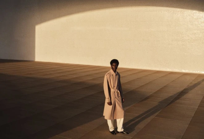 Model standing on a tiled surface wearing a beige trench coat under dramatic lighting for H&M