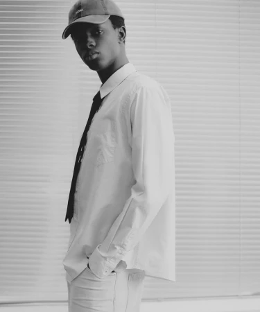 Monochrome portrait of a young man wearing a cap, white shirt, and tie against a horizontal slatted blind background.