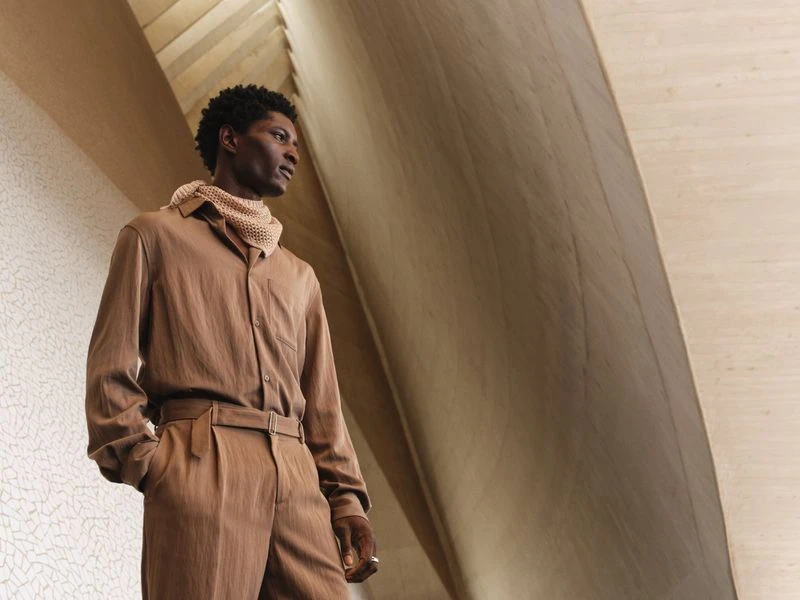 Adult male in neutral brown shirt and pants looking sideways in architectural interior with neutral textured wall in soft diffused light.