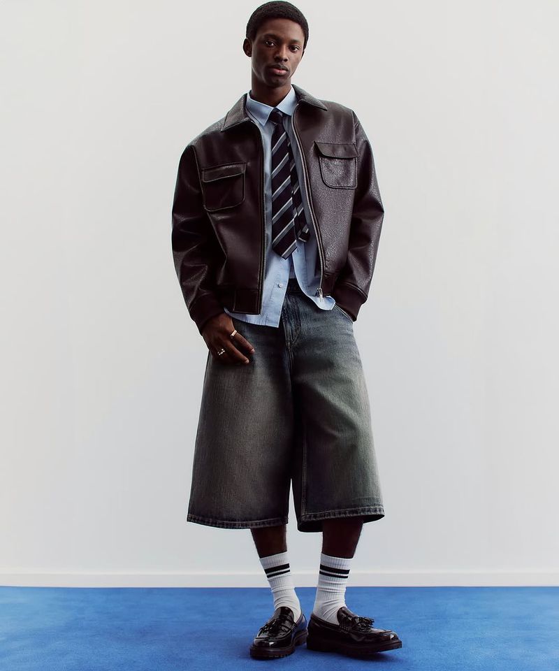Fashionable adult male in leather jacket, blue shorts, blue shirt, and black loafers posing in a light-filled photo studio.
