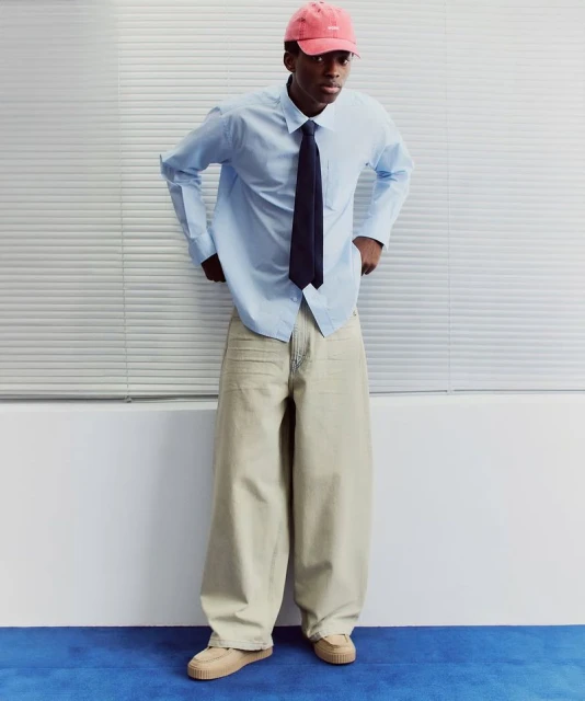 An African American man models light blue shirt, dark tie, baggy jeans, and pink cap against a white background with blue floor. 