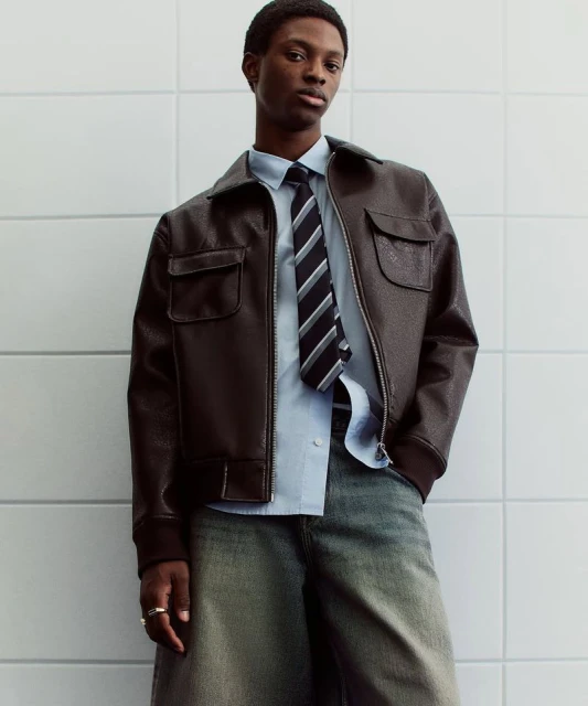 Adult male fashion model in a dark brown leather jacket, light blue shirt, and striped tie posing in front of a white tiled wall.