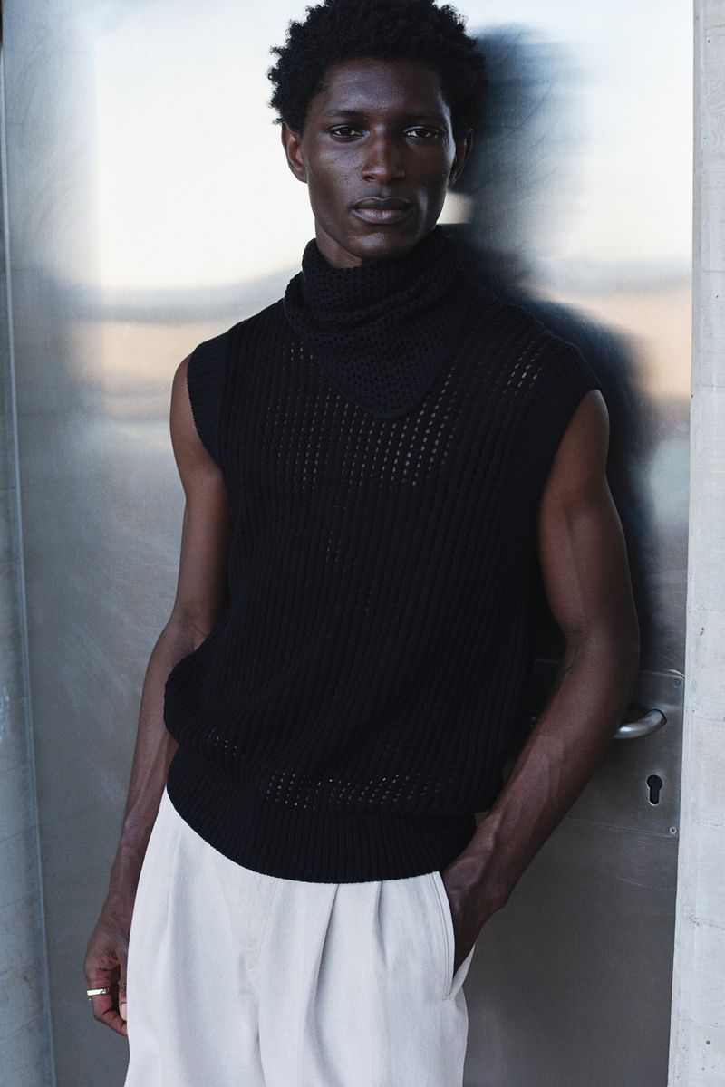 Fashionable portrait of a Black male model wearing a black knit sweater vest and beige pants, posing against a grey metallic wall.