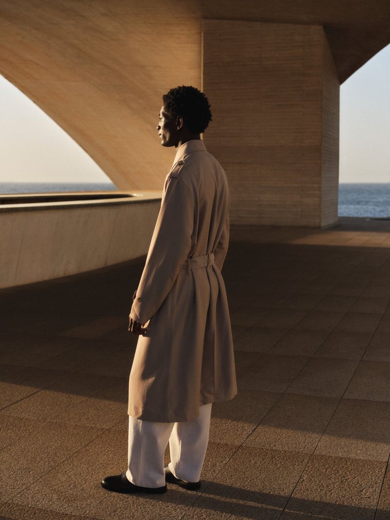 Model standing outdoors wearing a long beige trench coat and white trousers beneath modern concrete architecture.