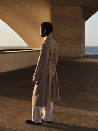 Model standing outdoors wearing a long beige trench coat and white trousers beneath modern concrete architecture.