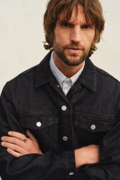 Man wearing a dark denim jacket and striped shirt with arms crossed against a pale background for H&M Spring 2025 Menswear.
