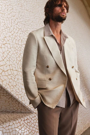 Man wearing a cream double-breasted blazer and striped shirt against a mosaic tile wall