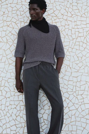 Black man wearing grey knit sweater and dark trousers leaning against a white mosaic tile wall