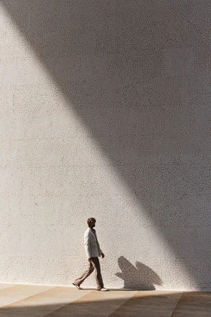 A man in a light jacket and trousers walking past a large textured wall with strong diagonal sunlight.