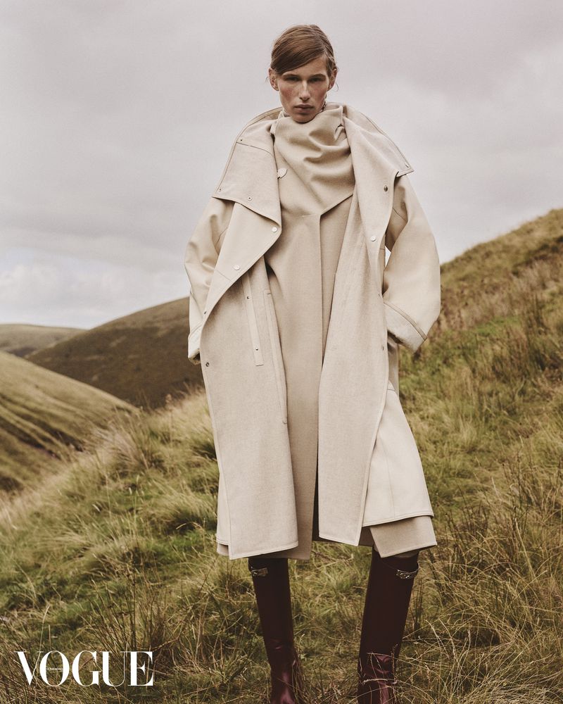 Fashion editorial shot of a single adult woman in an oversized beige trench coat and dark boots, standing in a grassy field with rolling hills.