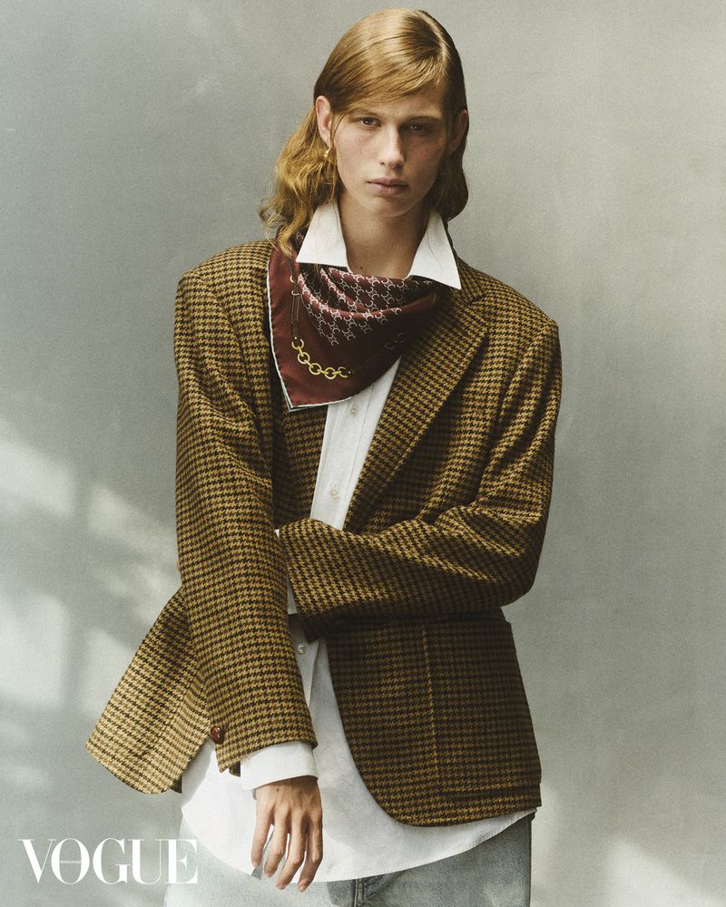 Adult male with medium-length brown hair poses in a brown tweed blazer, white shirt and silk scarf against a neutral backdrop.