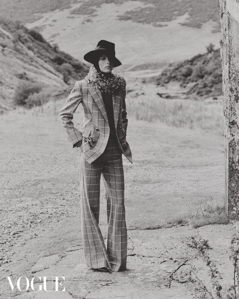 A woman poses outdoors in a black and white fashion shot, wearing a plaid suit, wide-brimmed hat, and fur scarf against a mountain backdrop.