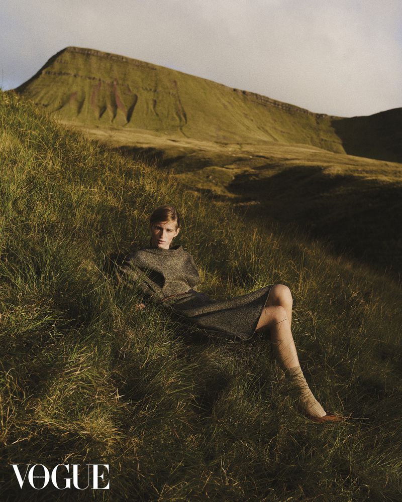 A woman in a grey dress and socks laying in a grassy field with a large mountain in the background under a cloudy sky.
