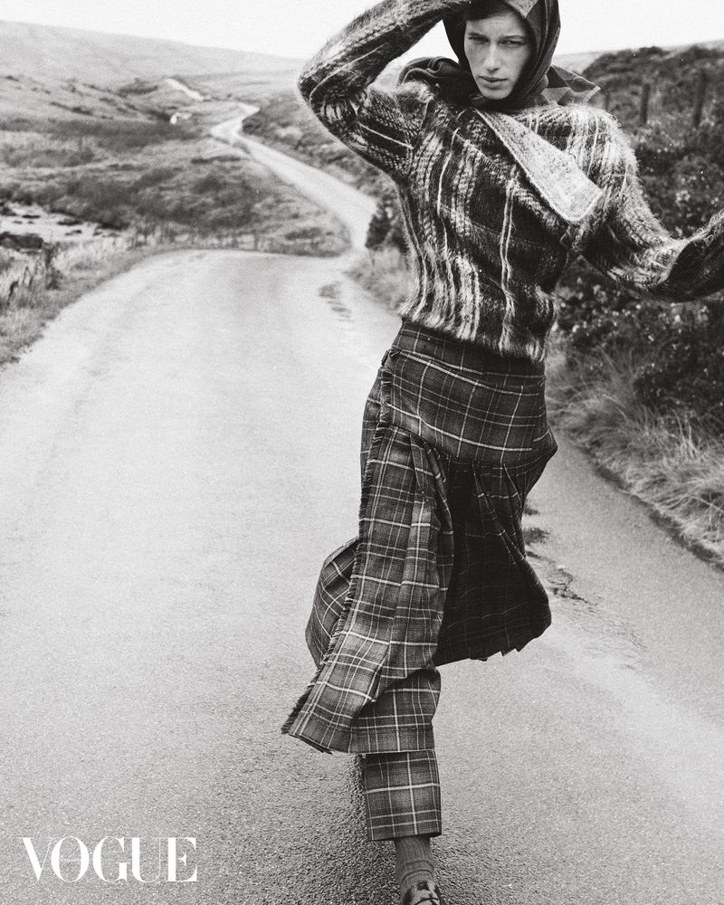 Model Esme Cornelius walking down a rural winding road wearing a plaid sweater, layered tartan skirt, and matching headscarf in monochrome.
