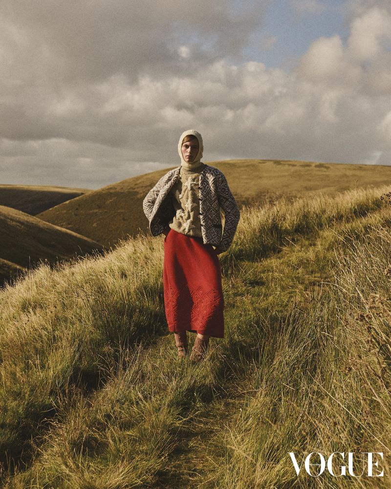 Woman in patterned jacket and red skirt stands on grassy hillside, framed by rolling hills and a cloudy sky, fashion editorial