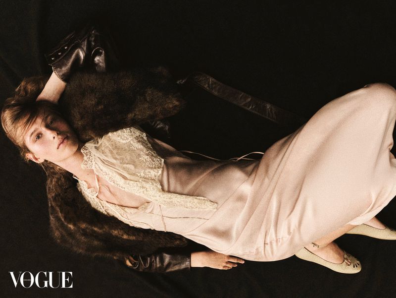 Elegant female model in a beige dress with lace details lying down on a black surface with leather and fur accents in a photo studio.