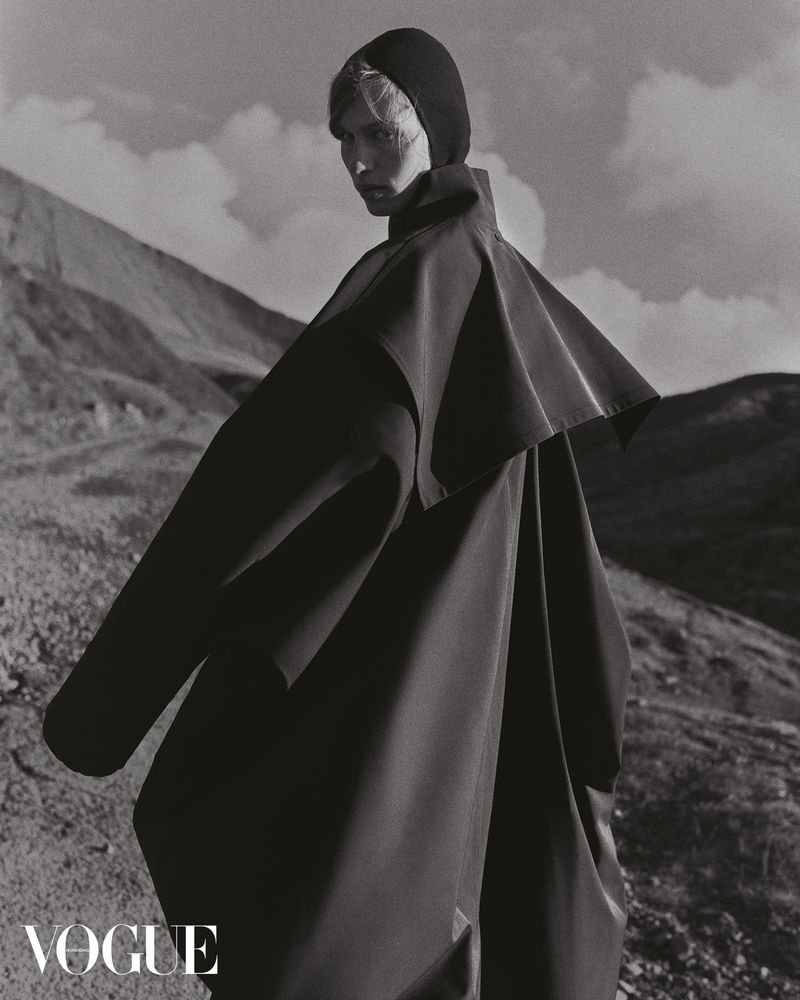 Monochrome fashion shot: a single adult poses in a large black hooded coat in a sparse, outdoor setting with rolling hills and cloudy sky.