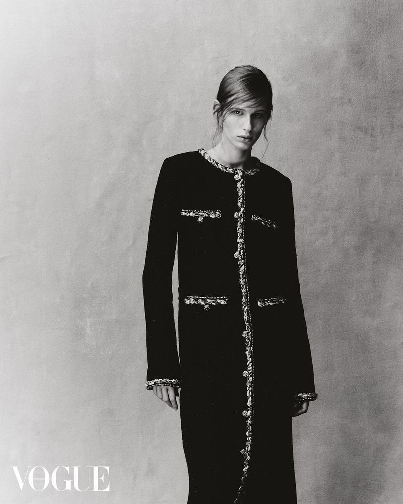 Monochrome fashion shot of a woman wearing a black jacket dress with decorative trim, posing against a textured grey backdrop. Vogue logo visible.