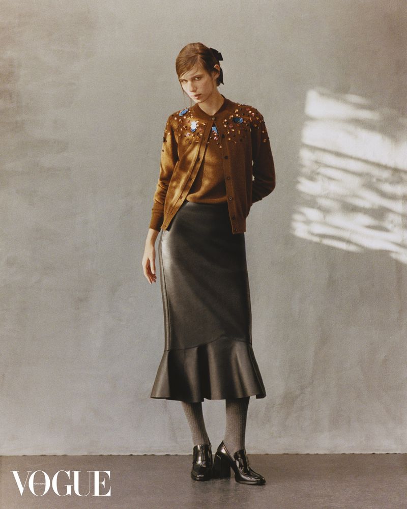 Woman in brown cardigan and black skirt, posing in front of a grey wall, fashion photography, full shot, neutral expression.