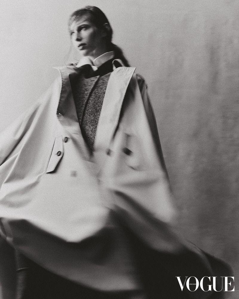 Monochrome studio fashion shot of a blonde woman in a large trench coat, styled with a dark sweater and collared shirt, posing for Vogue.