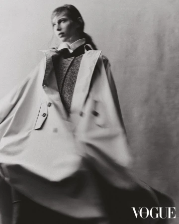 Monochrome studio fashion shot of a blonde woman in a large trench coat, styled with a dark sweater and collared shirt, posing for Vogue.
