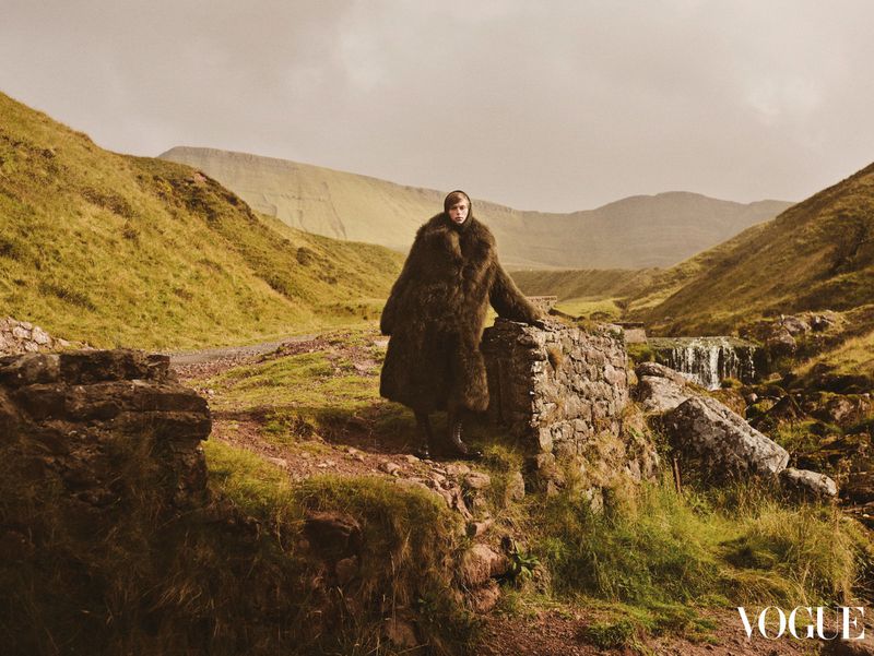 Woman in a fur coat stands on a stone wall with a scenic view of mountains and a small waterfall in the background, editorial fashion.
