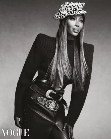 Naomi Campbell poses in a structured black knit top, beaded cap, and ornate western belts against a studio backdrop.