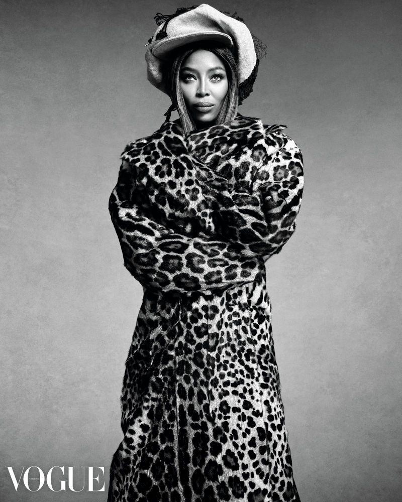 Monochrome portrait of Naomi Campbell wearing an animal-print coat and a hat against a gray backdrop, captured in a studio setting.
