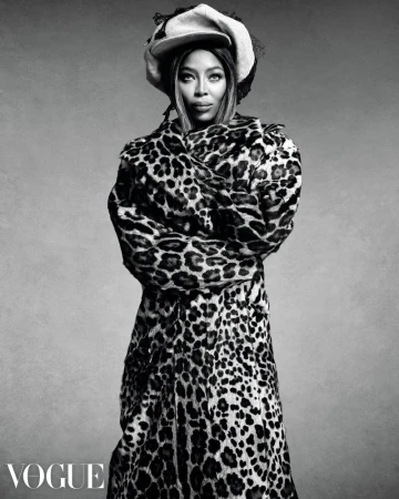 Monochrome portrait of Naomi Campbell wearing an animal-print coat and a hat against a gray backdrop, captured in a studio setting.