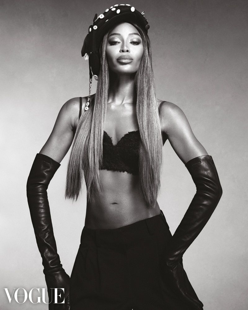 Black and white portrait of a woman with long straight hair wearing a hat, gloves, pants, and a bra, posing in a photo studio.