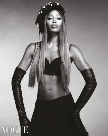 Black and white portrait of a woman with long straight hair wearing a hat, gloves, pants, and a bra, posing in a photo studio.