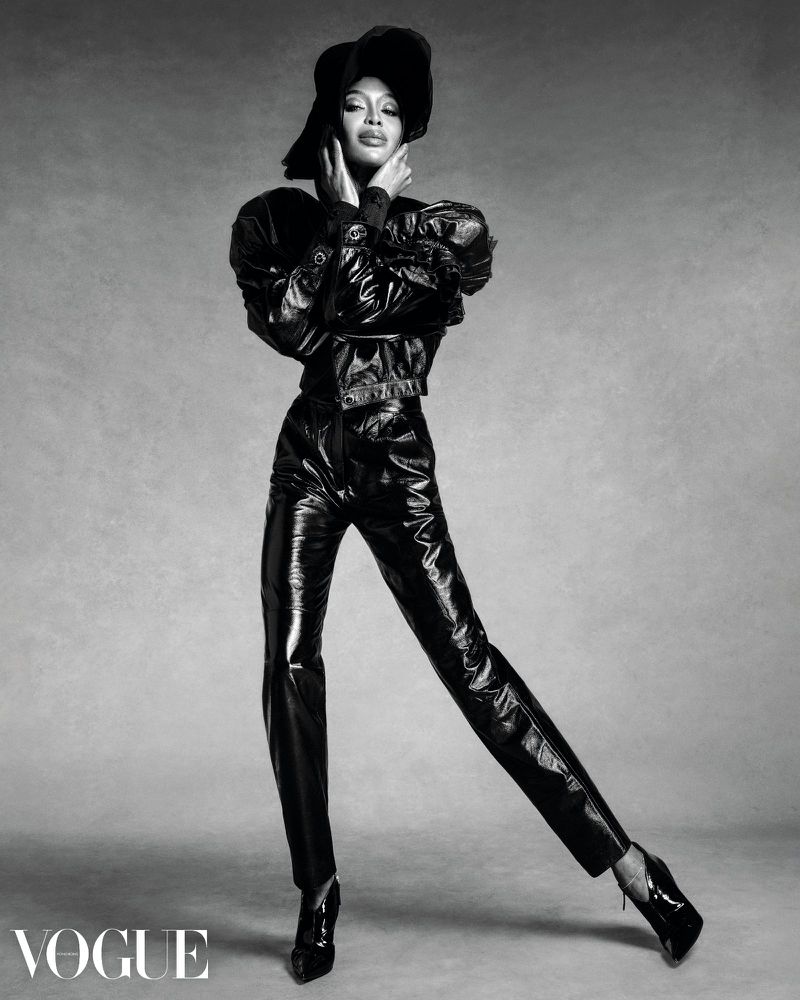 Black and white fashion shot of a Black woman in a hat, jacket, and pants, posing against a grey backdrop in a studio lighting.
