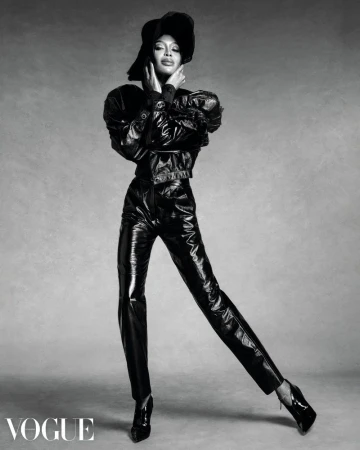 Black and white fashion shot of a Black woman in a hat, jacket, and pants, posing against a grey backdrop in a studio lighting.