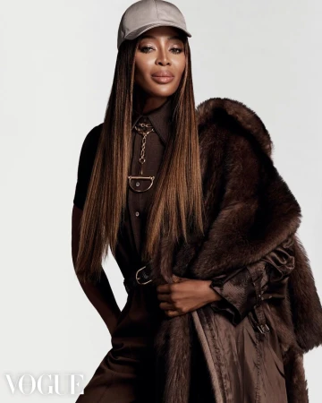 Brunette woman with tan skin in a brown jumpsuit, fur coat, and baseball cap poses against a white backdrop in a medium shot.