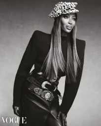 Naomi Campbell posing in a purple coat, sheer violet lace top, and green skirt against a white studio background.