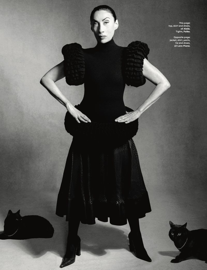 Woman in black dress with ruffled sleeves poses with two black cats in a studio, creating a high-fashion editorial shot.