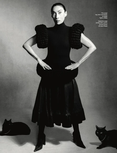 Woman in black dress with ruffled sleeves poses with two black cats in a studio, creating a high-fashion editorial shot.