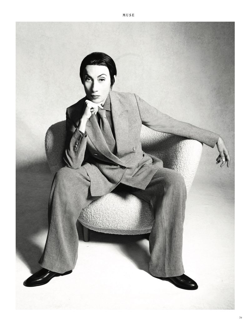 Monochrome portrait of a person in a suit sitting in a white chair, with a hand resting on their chin, and gazing directly at the camera.