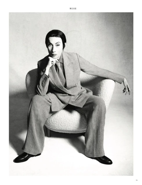 Monochrome portrait of a person in a suit sitting in a white chair, with a hand resting on their chin, and gazing directly at the camera.