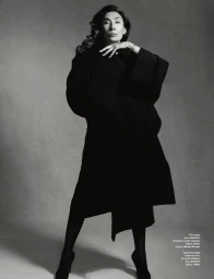 Monochrome fashion portrait of an older woman in a long black coat against a neutral studio background.