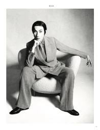 Model seated in a white textured armchair wearing a grey tailored suit against a neutral studio background.