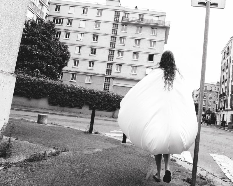 Person wearing a voluminous white garment walks down a city street, framed by urban buildings and trees in black and white.