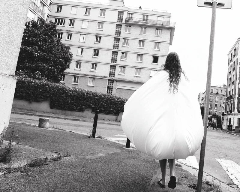Person wearing a voluminous white garment walks down a city street, framed by urban buildings and trees in black and white.